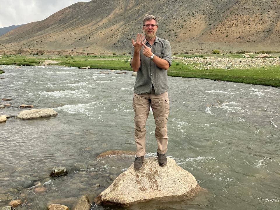 Taylor smiling and pointing to a ring on his finger while standing on a rock in a river surrounded by mountains